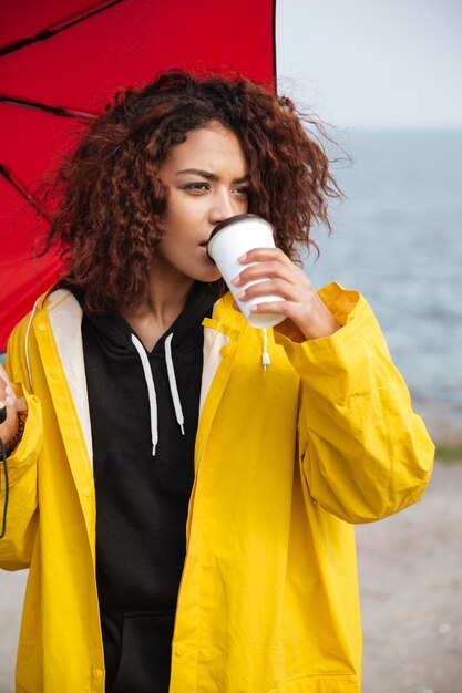 Konzentrierte afrikanische lockige junge Dame, die draußen geht und Kaffee trinkt.