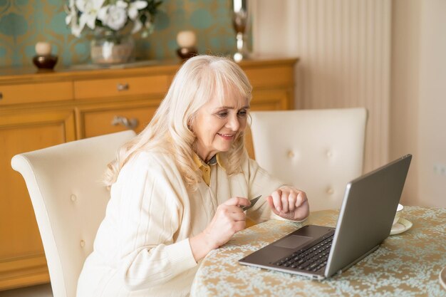 Konzentrierte alte Dame hält eine Kreditkarte vor einem schicken Laptop