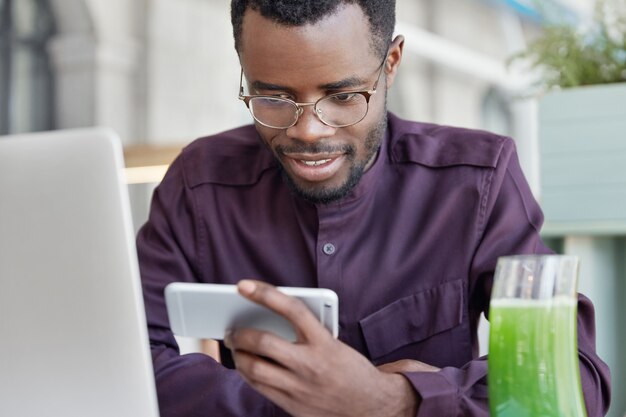 Konzentrierte begeisterte afroamerikanische Studenten schauen sich Filme oder Videos auf dem Smartphone an, tragen formelle Kleidung und eine runde Brille