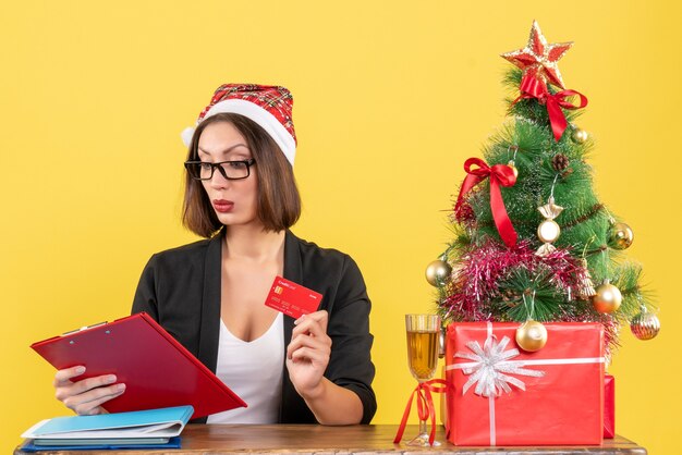 Konzentrierte charmante Dame im Anzug mit Weihnachtsmannhut und Brille, die Bankkarte im Büro auf gelb lokalisiert zeigt