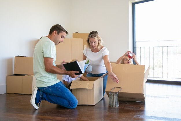 Konzentrierte Eltern und lustige Kinder packen Dinge in einer neuen Wohnung aus, sitzen auf dem Boden und nehmen Gegenstände aus der offenen Kiste