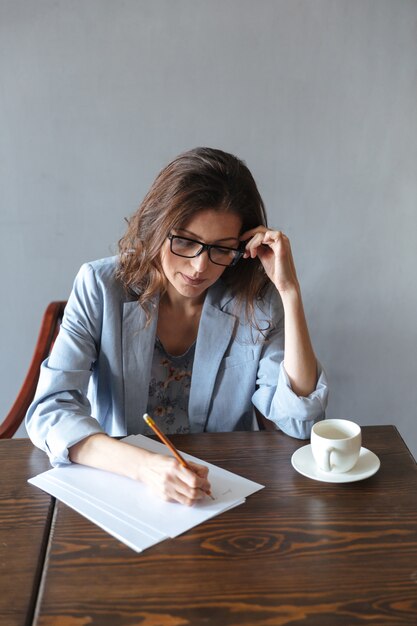 Konzentrierte Frau, die Notizen drinnen nahe Tasse Kaffee schreibt