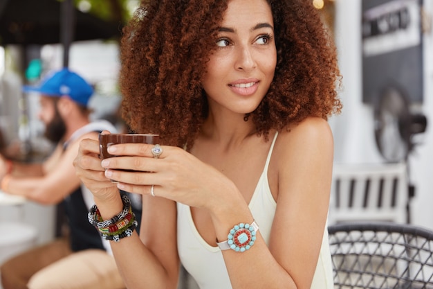 Konzentrierte Frau mit Afro-Frisur, genießt Kaffeepause in der Cafeteria, bemerkt jemanden hinter sich, schaut nachdenklich weg.