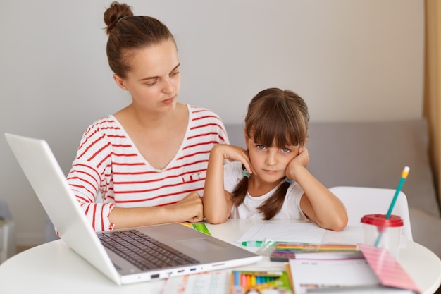 Konzentrierte Frau sitzt mit ihrer Tochter am Tisch mit Büchern und Laptop, macht Hausaufgaben oder hat Online-Unterricht, Mutter hilft ihrem Kind, Fernunterricht.