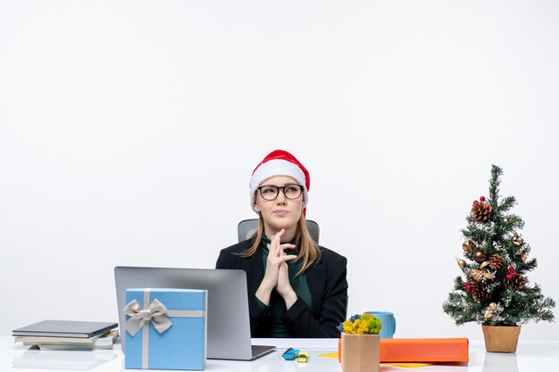 Konzentrierte Geschäftsfrau mit Weihnachtsmannhut, der an einem Tisch mit einem Weihnachtsbaum und einem Geschenk darauf im Büro auf weißem Hintergrund sitzt