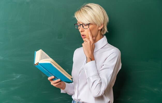 Konzentrierte junge blonde Lehrerin mit Brille im Klassenzimmer, die in der Profilansicht vor der Tafel steht, die ein Buch hält und liest und die Hand am Kinn hält