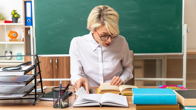 konzentrierte junge blonde Lehrerin mit Brille sitzt am Schreibtisch mit Schulwerkzeugen im Klassenzimmer und greift sich Notizblock, der die Hand am offenen Buch hält und das Buch betrachtet