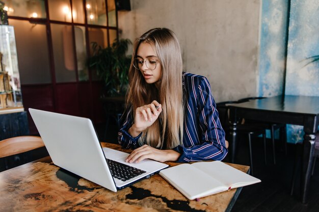 Konzentrierte junge Frau, die im Lieblingscafé arbeitet. Innenaufnahme der bezaubernden ernsten Studentin unter Verwendung des Laptops.