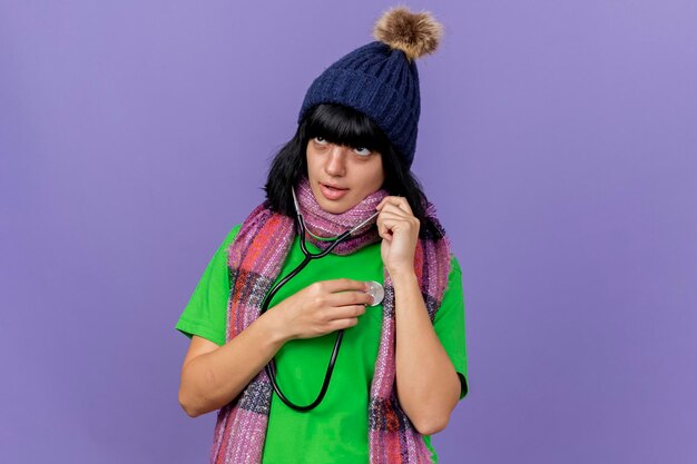 Konzentrierte junge kranke Frau, die Wintermütze und Schal mit Stethoskop trägt, das Seite betrachtet, der ihren Herzschlag lokalisiert auf purpurroter Wand mit Kopienraum hört