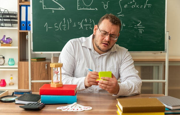 Kostenloses Foto konzentrierte junge lehrerin mit brille, die am schreibtisch mit schulmaterial im klassenzimmer sitzt und mit stift auf papiernotizen schreibt