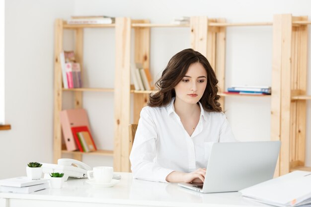 Konzentrierte junge schöne Geschäftsfrau, die an Laptop und Dokument in einem hellen, modernen Büro arbeitet