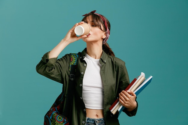 Konzentrierte junge Studentin mit Bandana und Rucksack mit Notizblöcken, die Kaffee aus einer Papierkaffeetasse trinken und in die Tasse schauen, die isoliert auf blauem Hintergrund steht