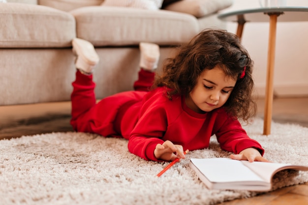 Konzentrierte Kinderzeichnung im Notizbuch. Innenaufnahme des niedlichen Kindes, das auf Teppich mit Stift liegt.
