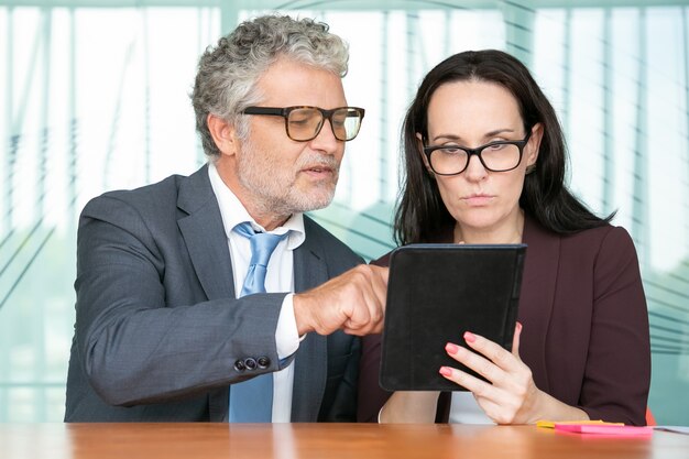 Konzentrierte Kollegen, die gemeinsam die Präsentation auf dem Tablet betrachten und auf den Bildschirm schauen, während sie im Büro am Tisch sitzen.