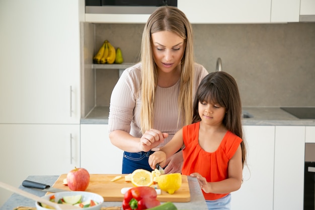 Konzentrierte Mutter beobachtet, wie ihr Mädchen frisches Gemüse im Schneidebrett schneidet. Kind hilft Mutter, Abendessen zu kochen. Familienkoch zusammen Konzept