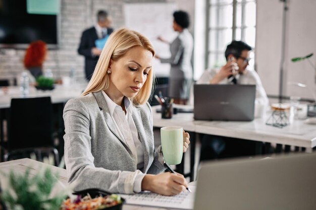 Konzentrierte Unternehmerin, die Notizen schreibt, während sie Geschäftsberichte im Büro analysiert. Im Hintergrund sind Menschen zu sehen