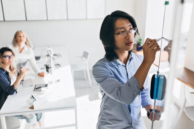 Konzentrierter asiatischer Manager, der Grafik während der Präsentation auf Flipchart zeichnet. Innenporträt des chinesischen Büroangestellten, der etwas auf weiße Tafel schreibt, während seine Kolleginnen zuschauen.