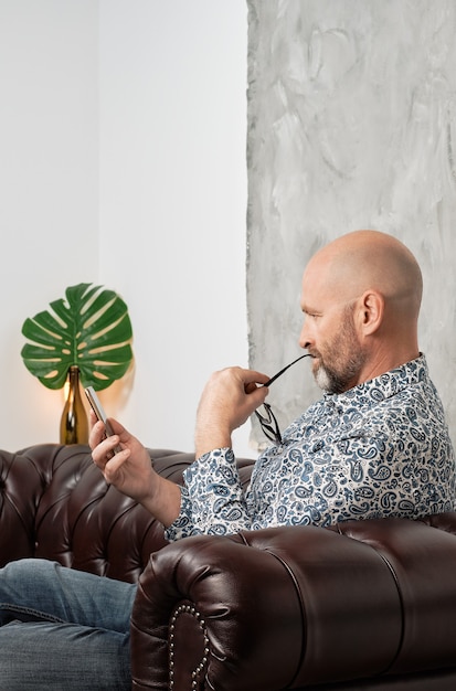 konzentrierter bärtiger Mann mit Brille und Smartphone in der Hand auf einem Stuhl sitzend