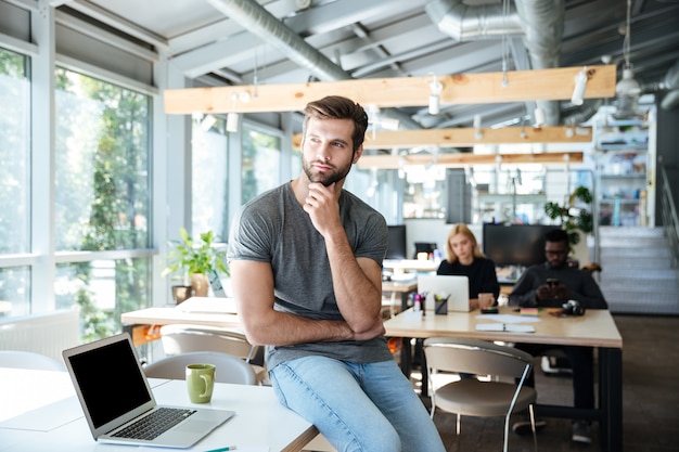 Konzentrierter denkender junger Mann, der auf Tisch im Büro sitzt