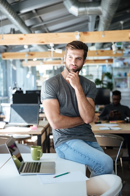 Konzentrierter denkender junger Mann, der auf Tisch im Büro sitzt