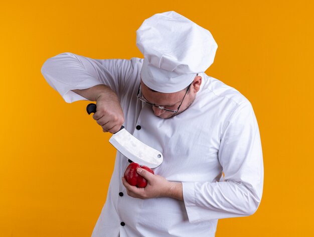 Konzentrierter erwachsener männlicher Koch mit Kochuniform und Brille, der Pfeffer mit Hackmesser berührt, der Pfeffer einzeln auf orangem Hintergrund betrachtet