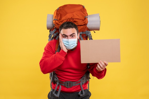 Konzentrierter fokussierter junger mann, der eine medizinische maske mit rucksack trägt und ein blatt hält, ohne auf isoliertem gelbem hintergrund zu schreiben writing