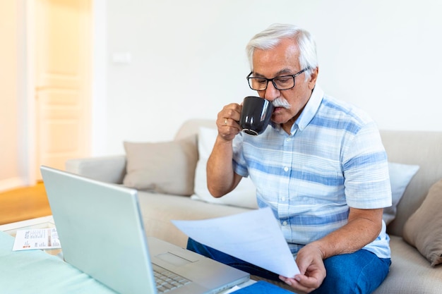 Konzentrierter grauhaariger älterer Mann sitzt auf der Couch und liest Bankbenachrichtigungen, die Haushaltsausgaben berechnen