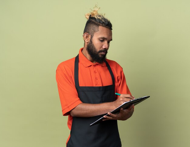 Konzentrierter junger afroamerikanischer Barbier, der eine Uniform trägt und die Zwischenablage mit Bleistift auf olivgrüner Wand mit Kopienraum schreibt