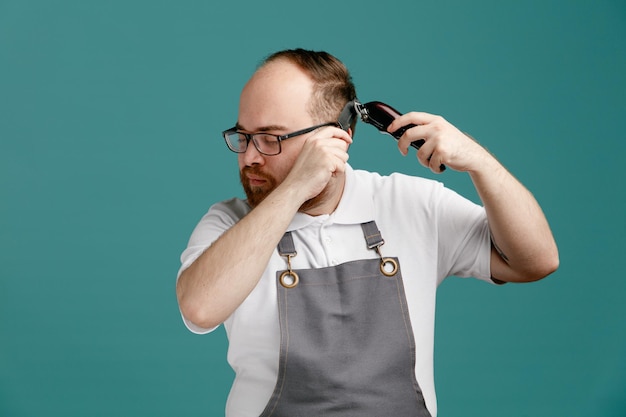 Konzentrierter junger Friseur in Uniform und Brille, der sich von Kopf zu Seite dreht und Teaserkamm und Haarschneider hält, der sein eigenes Haar mit geschlossenen Augen einzeln auf blauem Hintergrund schneidet