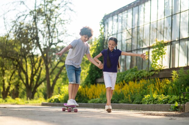 Konzentrierter junger Skateboard-Anfänger, der versucht, das Gleichgewicht auf dem Board zu halten, unterstützt von seinem Freund