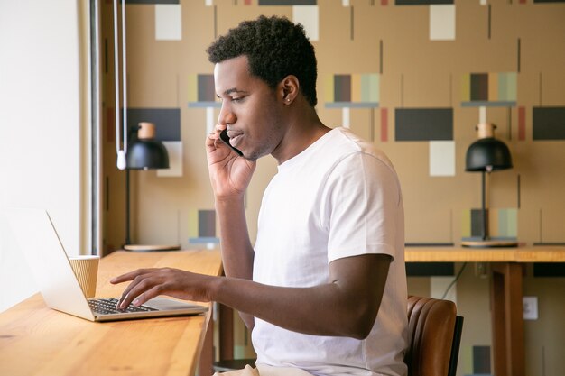 Konzentrierter junger Unternehmer, der am Laptop arbeitet und im gemeinsamen Arbeitsbereich auf dem Handy spricht