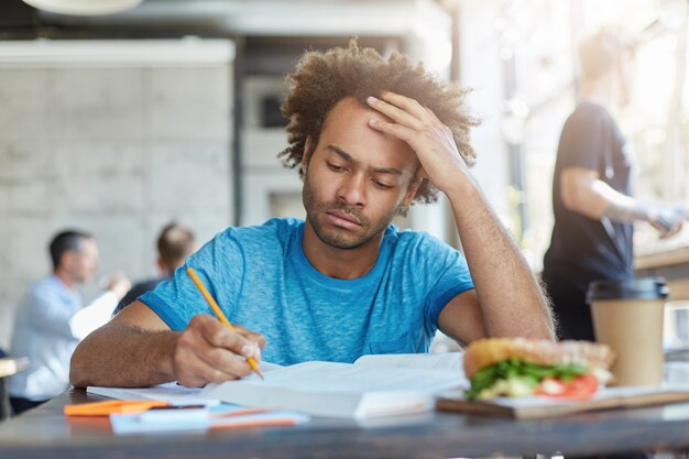 Konzentrierter, lässig gekleideter, dunkelhäutiger, unrasierter Student, der im Café studiert, in ein Heft schreibt, Nachforschungen anstellt oder sich auf die Prüfung am College vorbereitet und ernsthaft hinschaut