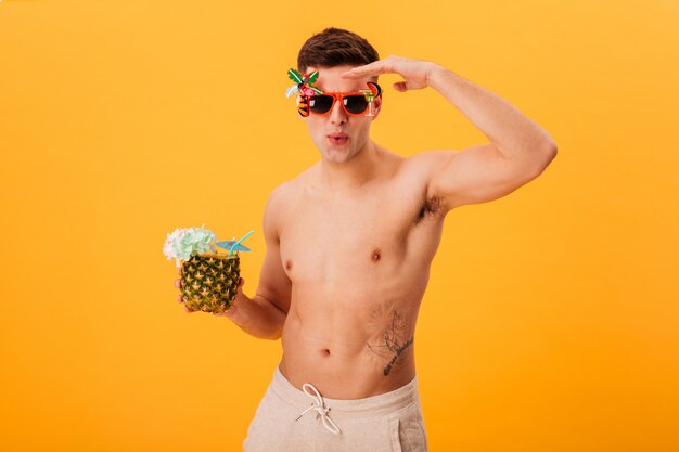 Konzentrierter nackter Mann in Shorts und ungewöhnlicher Sonnenbrille mit Cocktail