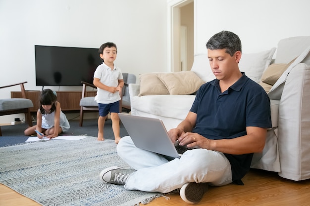 Konzentrierter Vater, der auf dem Boden sitzt und PC benutzt, während seine kleinen Kinder zeichnen und gehen