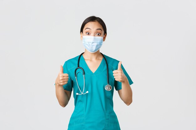 Konzept der Covid19-Coronavirus-Krankheit im Gesundheitswesen Beeindruckte, glückliche asiatische Ärztin in medizinischer Maske und Scrubs zeigen Daumen hoch, stimmen zu, Kompliment, Wahl, Lob, gute Arbeit