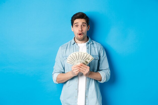 Konzept der Finanzen, Kredite und Banken. Überraschter Mann mit Geld, Blick in die Kamera wunderte sich und stand über blauem Hintergrund.