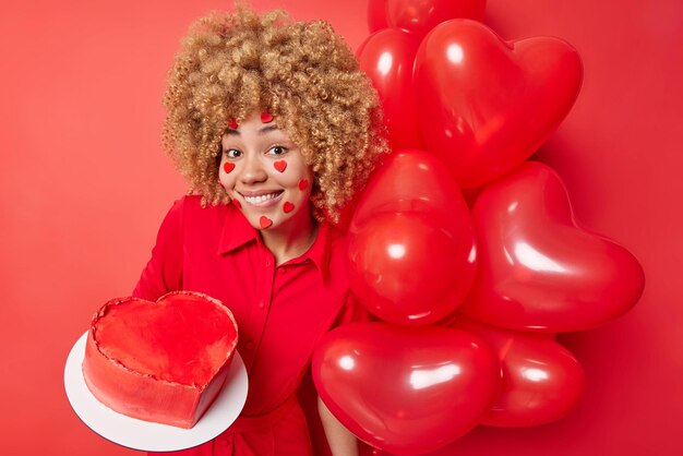 Konzept der Geburtstagsfeier Lächelnde Europäerin mit lockigen blonden Haaren trägt ein festliches Kleid, das gut gelaunt ist, hält Kuchen und einen Haufen aufgeblasener Luftballons isoliert über leuchtend rotem Hintergrund