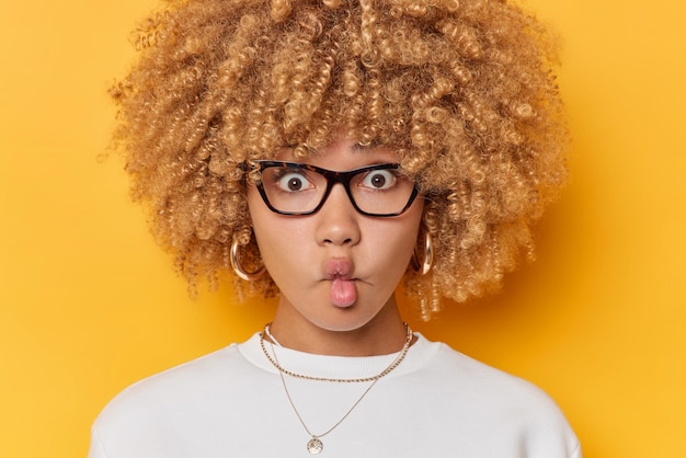 Kostenloses Foto konzept der menschlichen gesichtsausdrücke überrascht lockige frau macht fischlippen hat sich gewundert, dass der ausdruck durch eine transparente brille starrt und einen lässigen weißen pullover trägt, der über gelbem hintergrund isoliert ist