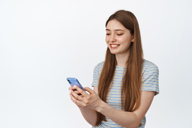 Konzept der Mobilfunktechnologie. Lächelnde junge Frau, die Handy-Scene mit zufriedenem Gesichtsausdruck betrachtet, weißer Hintergrund.