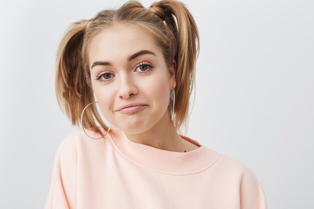 Konzept der modernen Generation, Jugend und Glück. Bild des lustigen stilvollen Studentenmädchens mit zwei Pferdeschwänzen, die ihre Freizeit drinnen verbringen, im rosa Sweatshirt und mit großen runden Ohrringen.