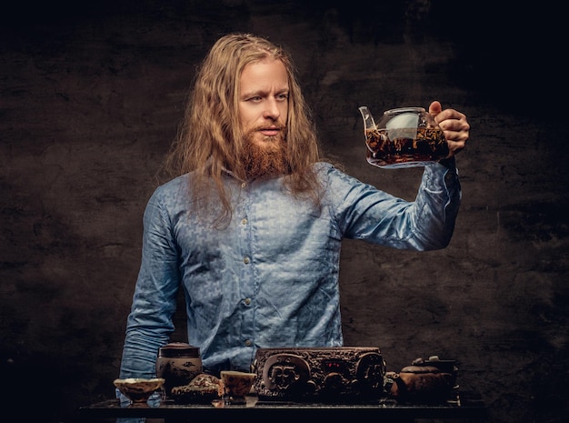 Konzept der Teezeremonie. Porträt eines rothaarigen Hipster-Männchens mit langen Haaren und Vollbart, gekleidet in ein blaues Hemd, konzentriert sich auf die Zubereitung von Tee mit einem handgefertigten Teeset. Getrennt auf einem dunklen strukturierten backgr