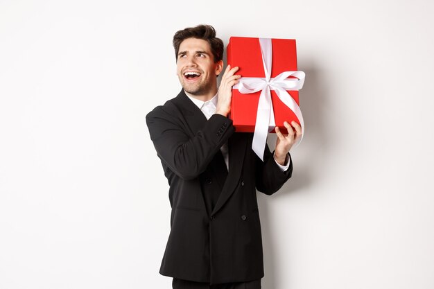 Konzept der Weihnachtsferien, Feiern und Lifestyle. Bild eines aufgeregten Mannes, der das neue Jahr genießt, Geschenkbox schütteln, um zu erraten, was drin ist und vor weißem Hintergrund steht.