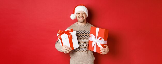 Konzept der Winterferien, des neuen Jahres und der Feier. Das Bild eines glücklichen und aufgeregten jungen Mannes mag Geschenke, hält Geschenke und lächelt, trägt eine Weihnachtsmütze und einen Weihnachtspullover