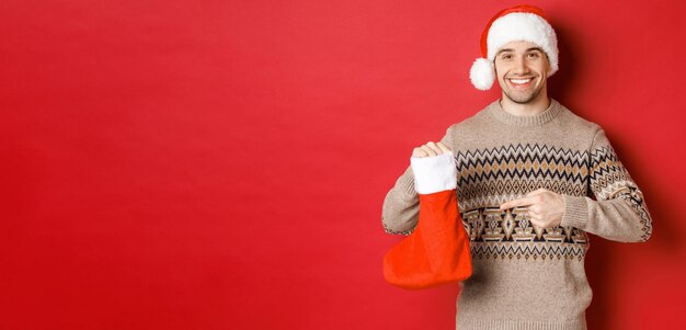 Konzept der Winterferien Neujahr und Feiern Gutaussehender lächelnder Mann bereitete Geschenke für Kinder vor, die auf die Weihnachtsstrumpftasche zeigen, die über rotem Hintergrund steht