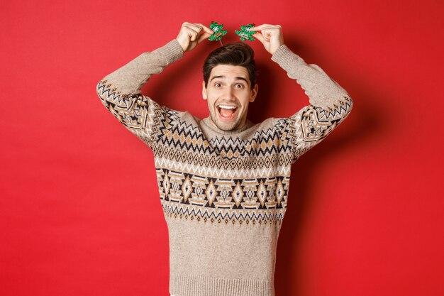Konzept der Winterferien, Weihnachten und Feiern. Bild eines lustigen und süßen erwachsenen Mannes, der die Neujahrsparty genießt, lächelt und sich freut und über rotem Hintergrund steht.
