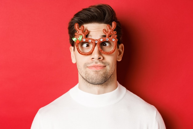 Konzept der Winterferien, Weihnachten und Feiern. Nahaufnahme der lustigen Brünette in Partybrille, schielen und Grimassen, stehend über rotem Hintergrund.