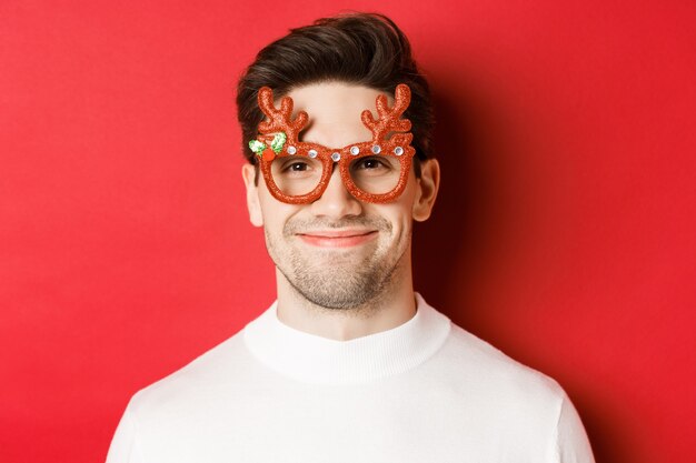 Konzept der Winterferien, Weihnachten und Feiern. Nahaufnahme des attraktiven lächelnden männlichen Modells in der Partybrille, stehend vor rotem Hintergrund