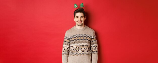 Konzept der Winterferien, Weihnachten und Feiern. Schöner bärtiger Kerl im Pullover, der ein frohes neues Jahr wünscht, in die Kamera lächelt und über rotem Hintergrund steht.