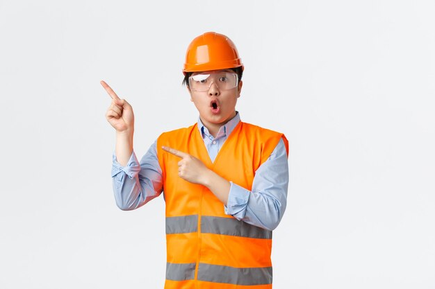 Konzept des Bausektors und der Industriearbeiter. Überraschter und beeindruckter asiatischer männlicher Ingenieur, Bauleiter in der Fabrik mit Schutzhelm, reflektierender Kleidung und zeigender oberer linker Ecke.