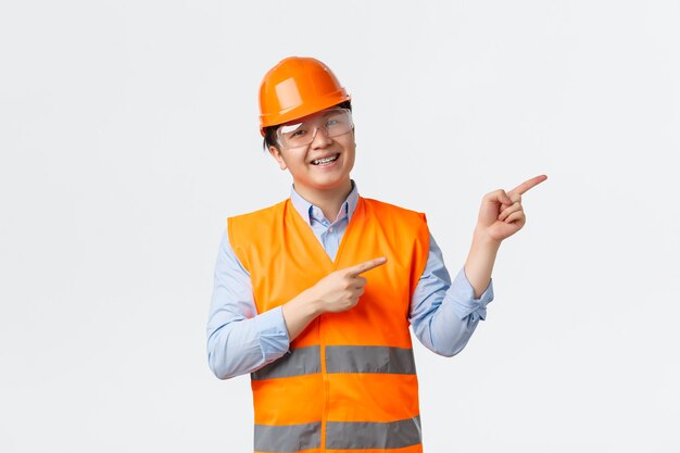 Konzept des Bausektors und der Industriearbeiter. Fröhlich lächelnder asiatischer Baumeister, Bauleiter in Helm und reflektierende Kleidung, die auf die obere rechte Ecke zeigt, demonstrieren, weißer Hintergrund.